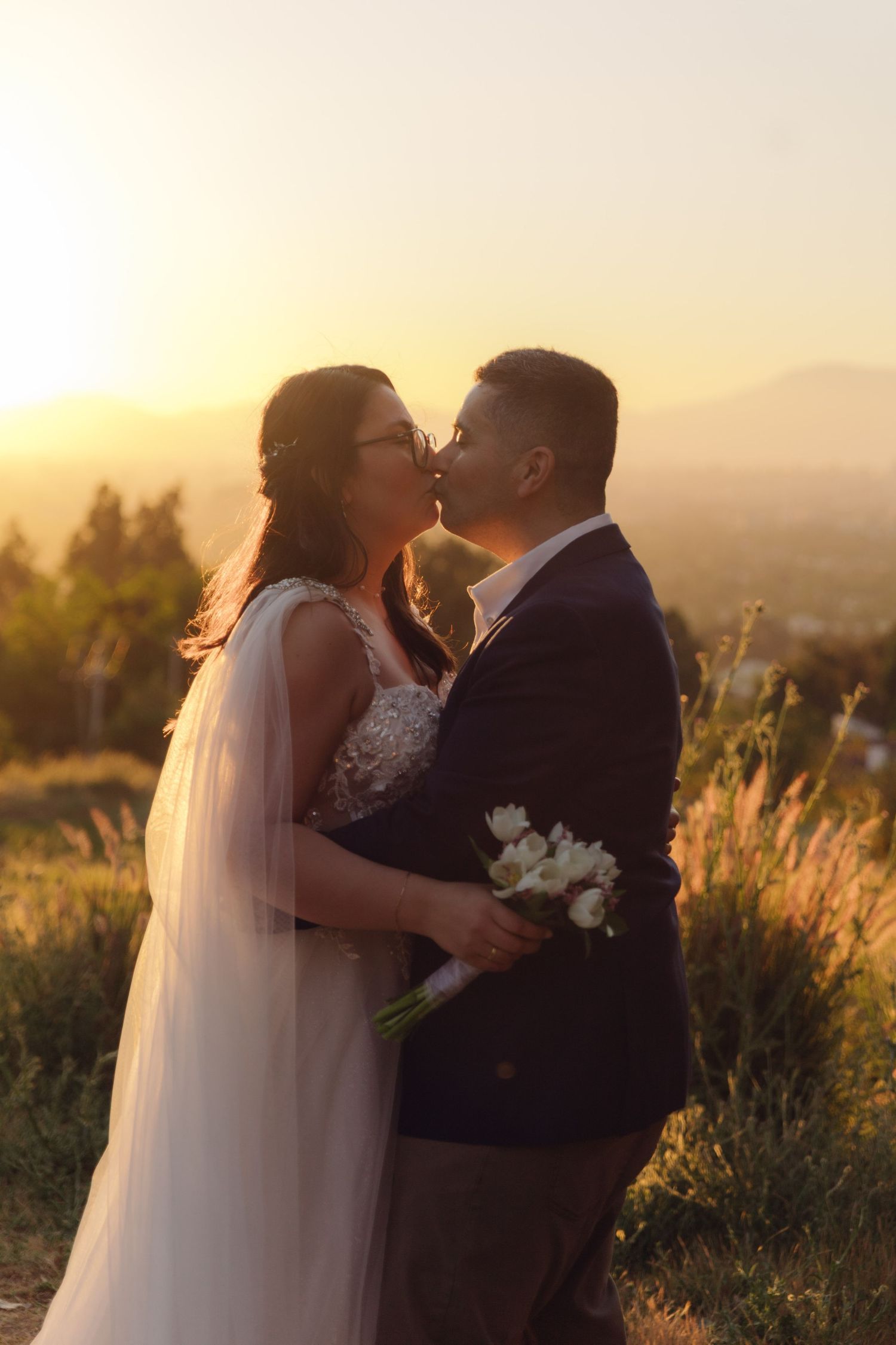 Newlyweds share a romantic kiss silhouetted against vibrant golden sunset with mountain valley views behind them.