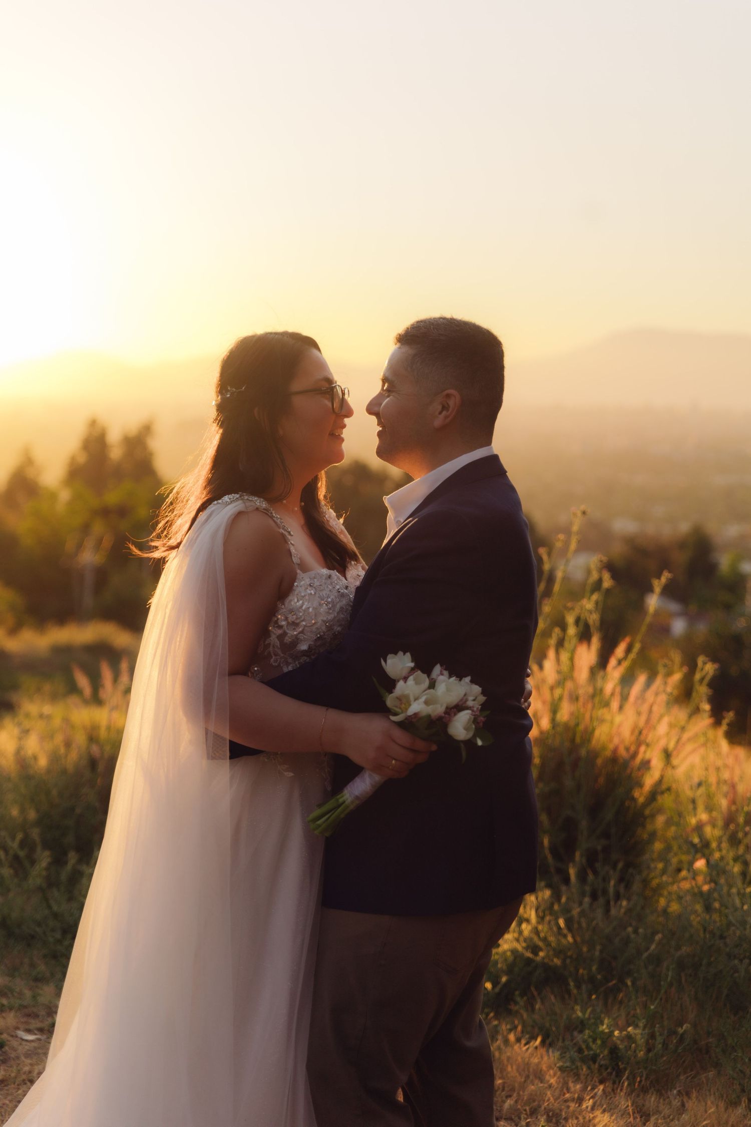 Bride in lace gown and groom in dark suit share tender moment together during breathtaking golden hour sunset.