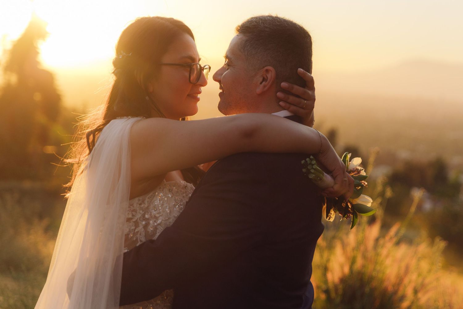 Bride embraces groom from behind in warm golden sunset light, holding bouquet while smiling softly together.