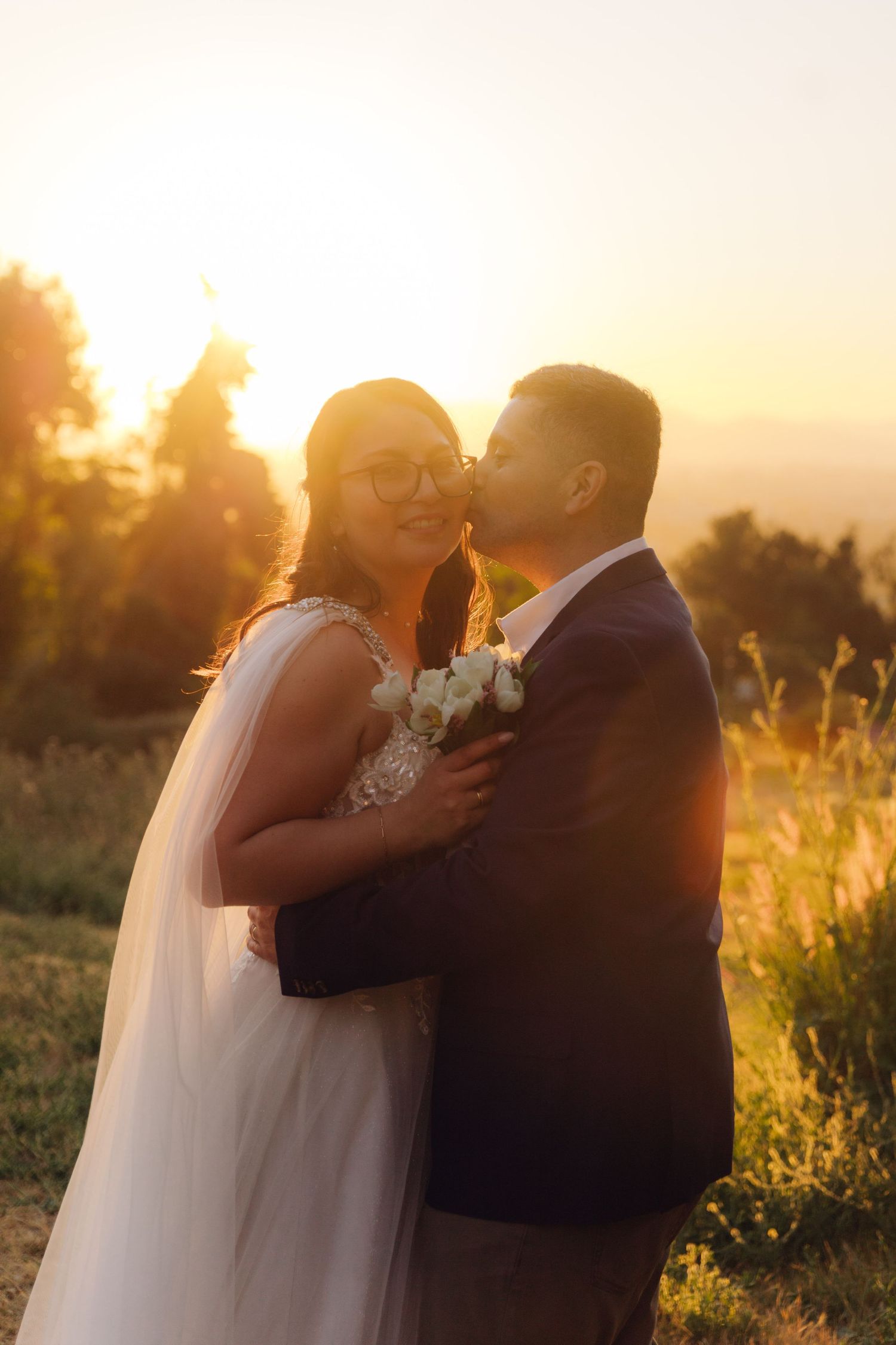 Groom kisses smiling bride on cheek during golden sunset, she holds white bouquet in romantic outdoor portrait.