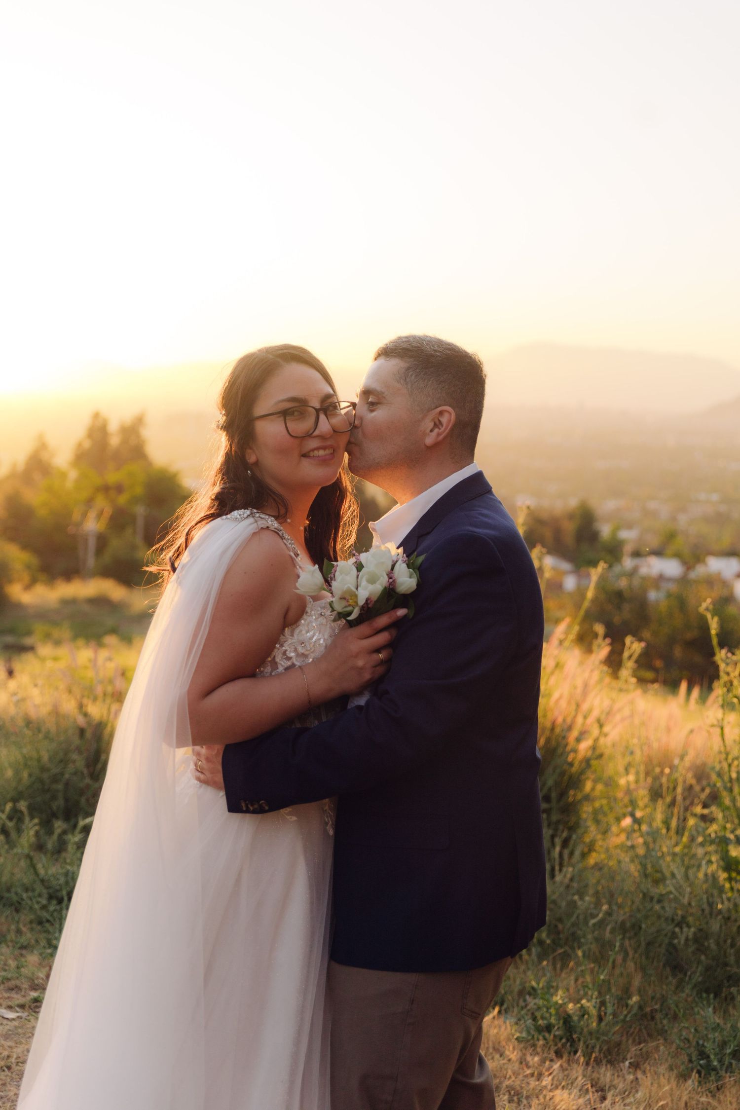 Groom tenderly kisses bride on cheek as she smiles, holding white roses during stunning golden hour sunset.
