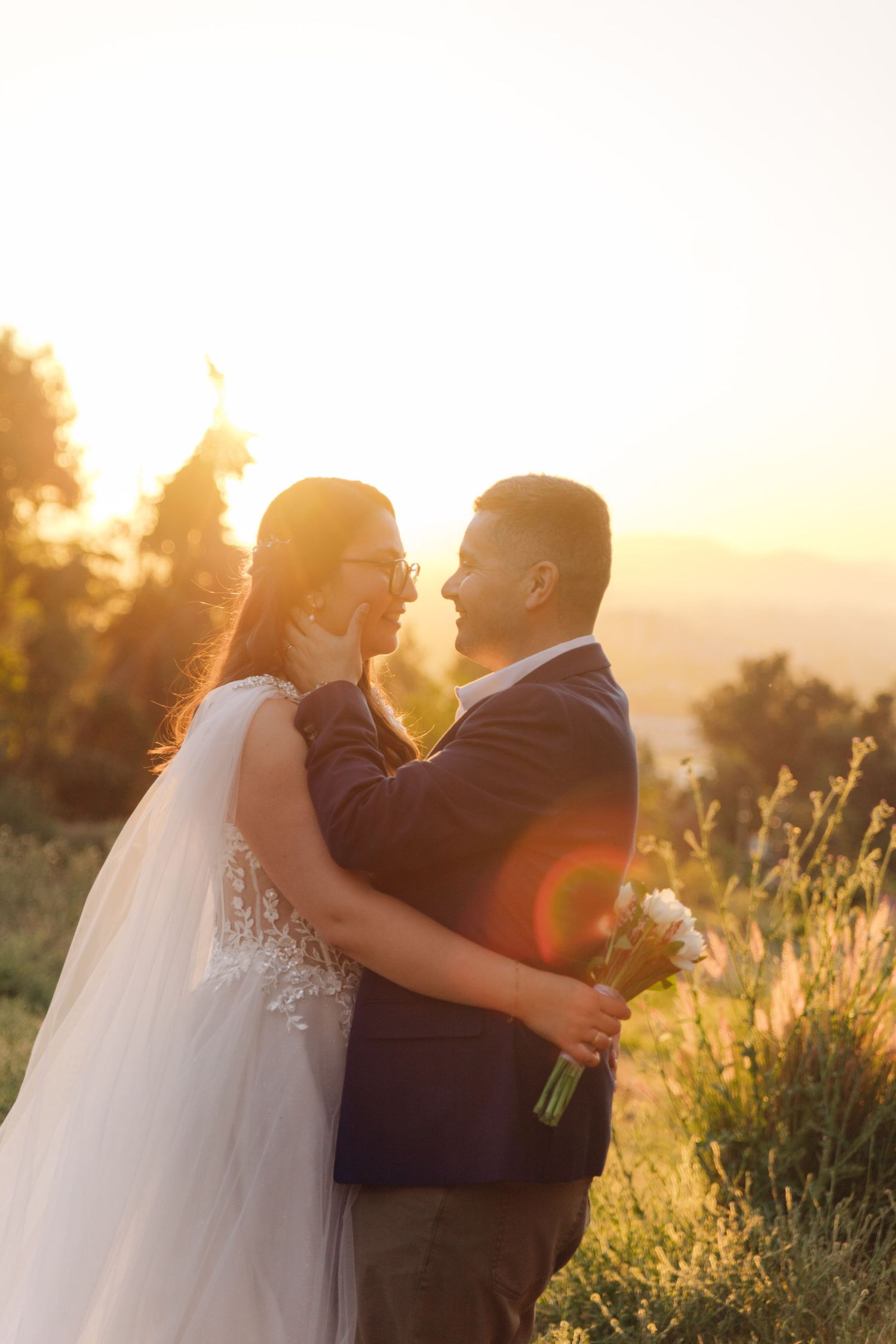 Bride and groom embrace at golden hour sunset with mountain views, bride holding white bouquet in wedding attire.