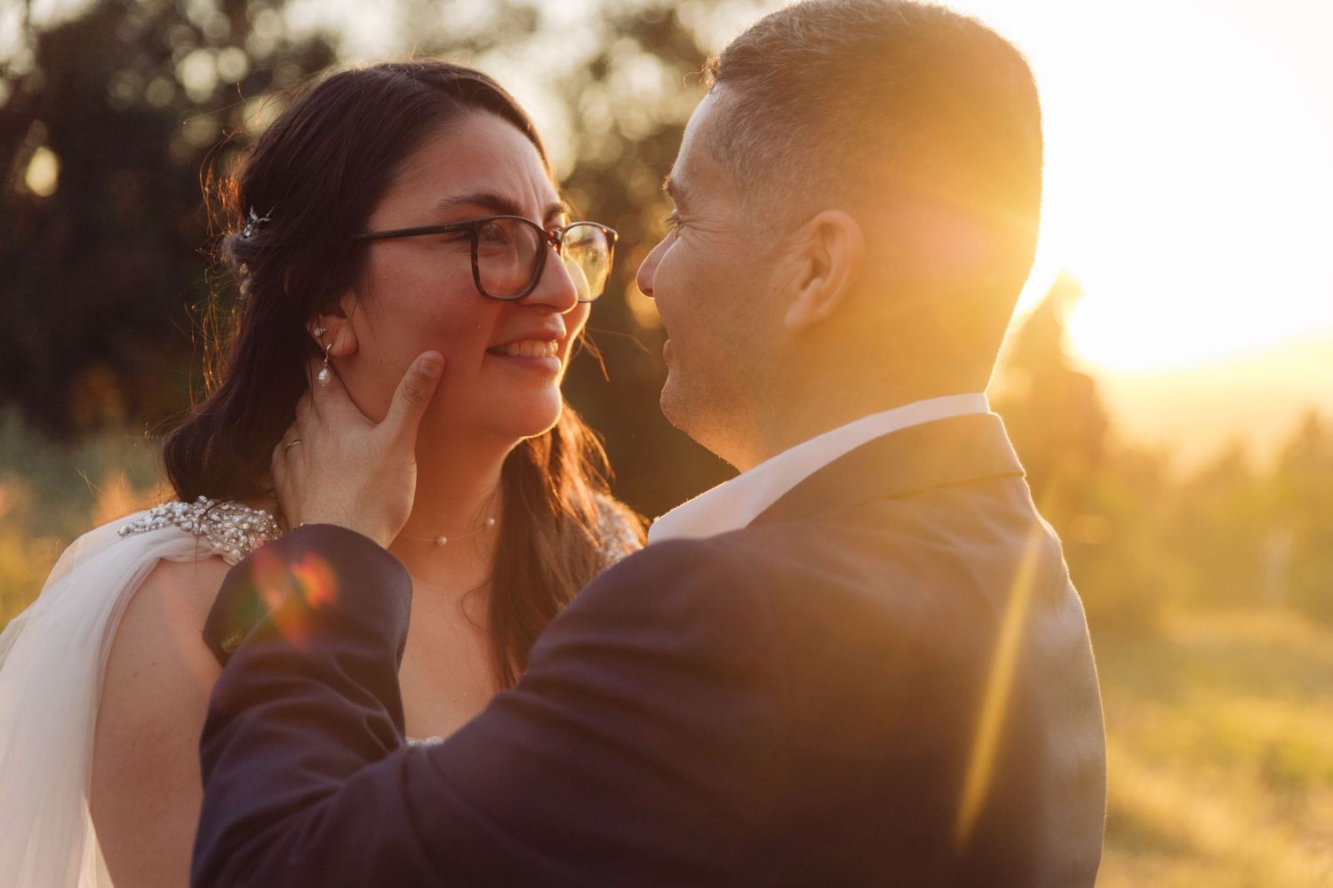 Groom tenderly cups brides face as she smiles wearing glasses, bathed in warm golden sunset light.