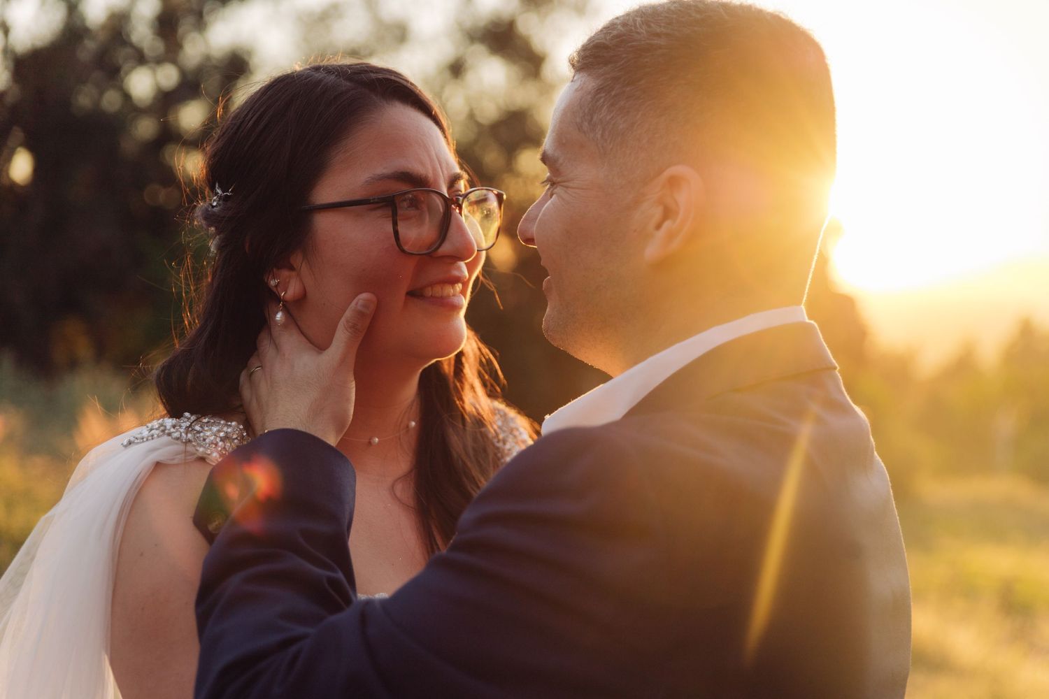 Close-up of groom holding brides face gently as she smiles in glasses during romantic sunset wedding shoot.