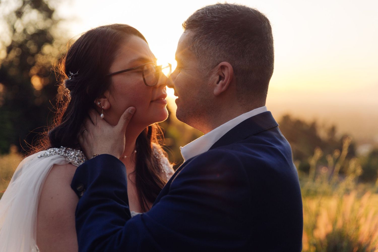 Bride and groom share an intimate almost-kiss moment backlit by glowing golden sunset light outdoors.