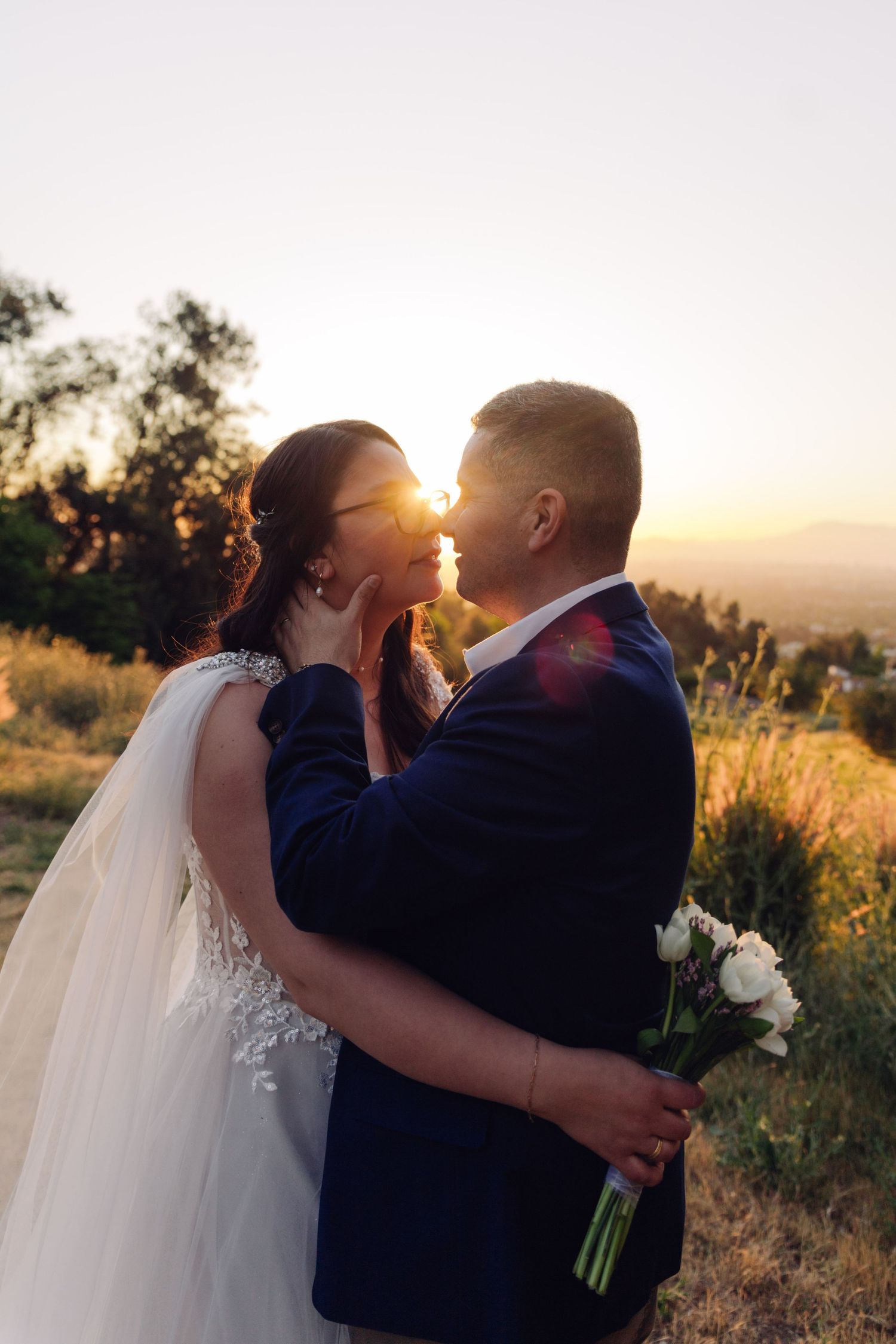 Wedding couple nearly kissing at sunset with bride in lace gown holding white bouquet in scenic outdoor setting.