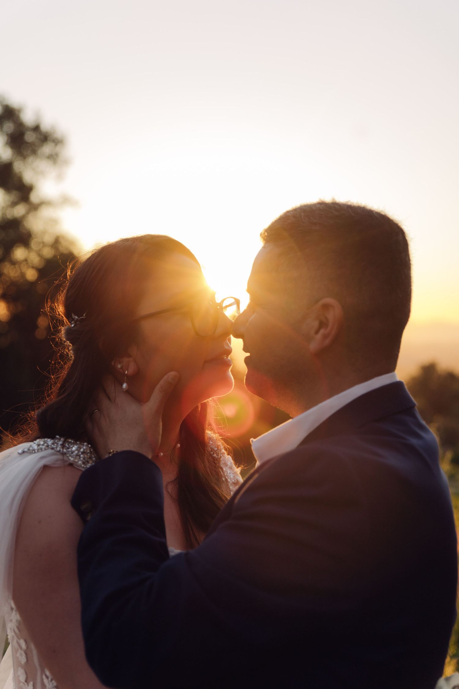 Bride and groom share a romantic kiss silhouetted against a vibrant golden sunset in an outdoor setting.