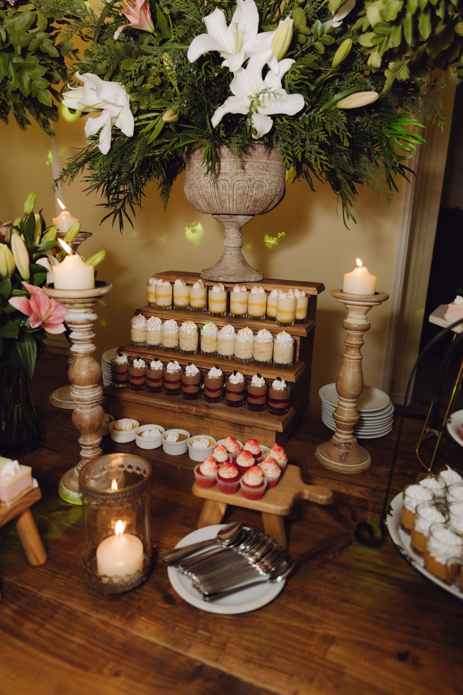 Rustic wedding dessert table with tiered mini cakes, candles, and large white lily floral arrangement centerpiece.