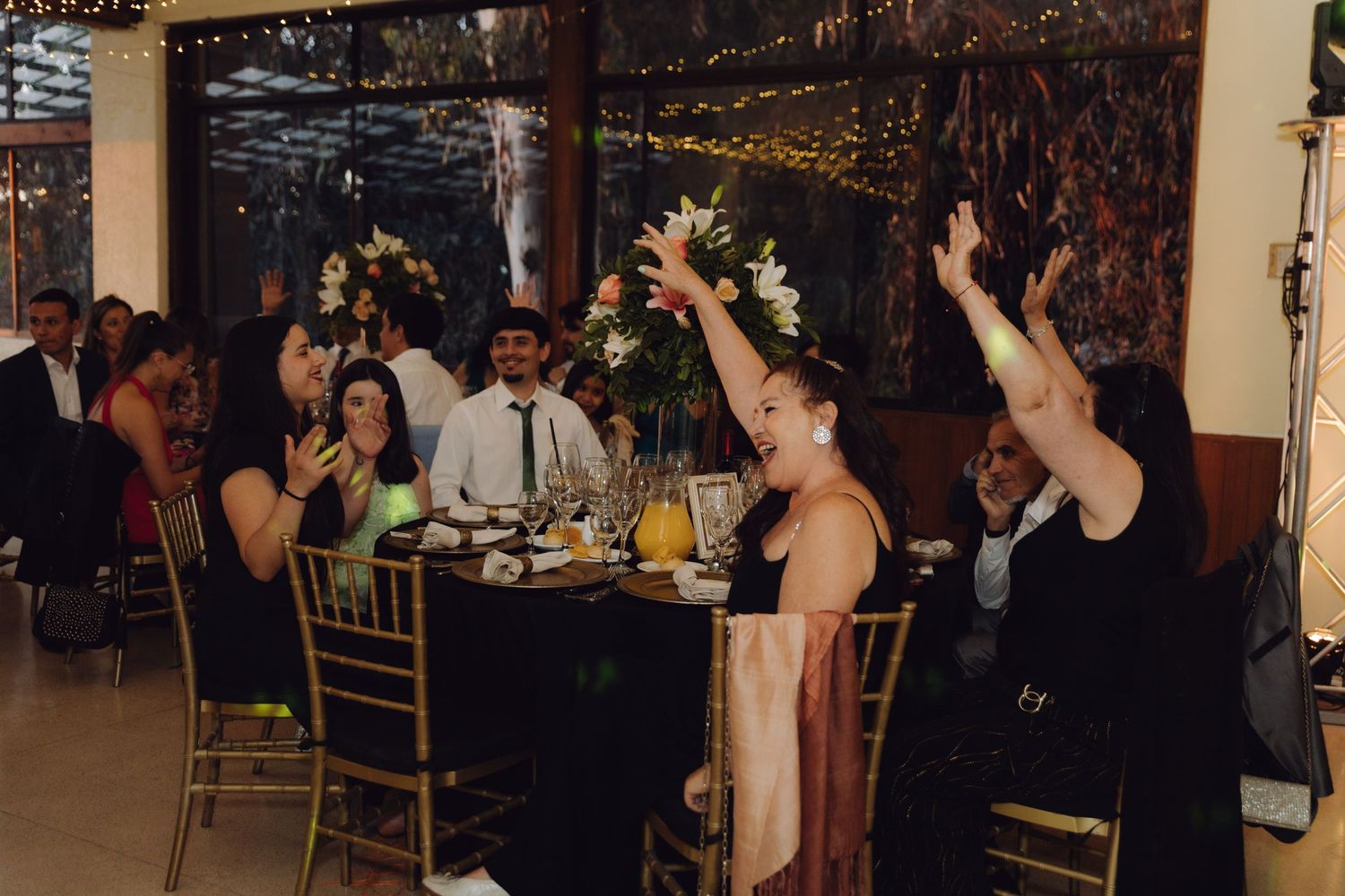 Joyful wedding reception guests celebrating and raising hands at candlelit tables with floral centerpieces.