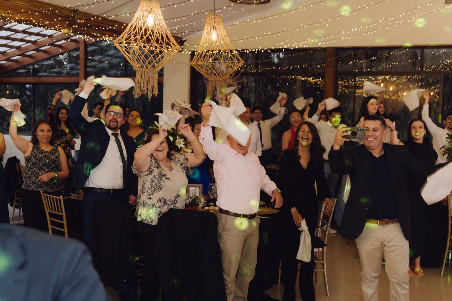 Joyful wedding reception guests waving white napkins and dancing under warm string lights and geometric pendant lamps.