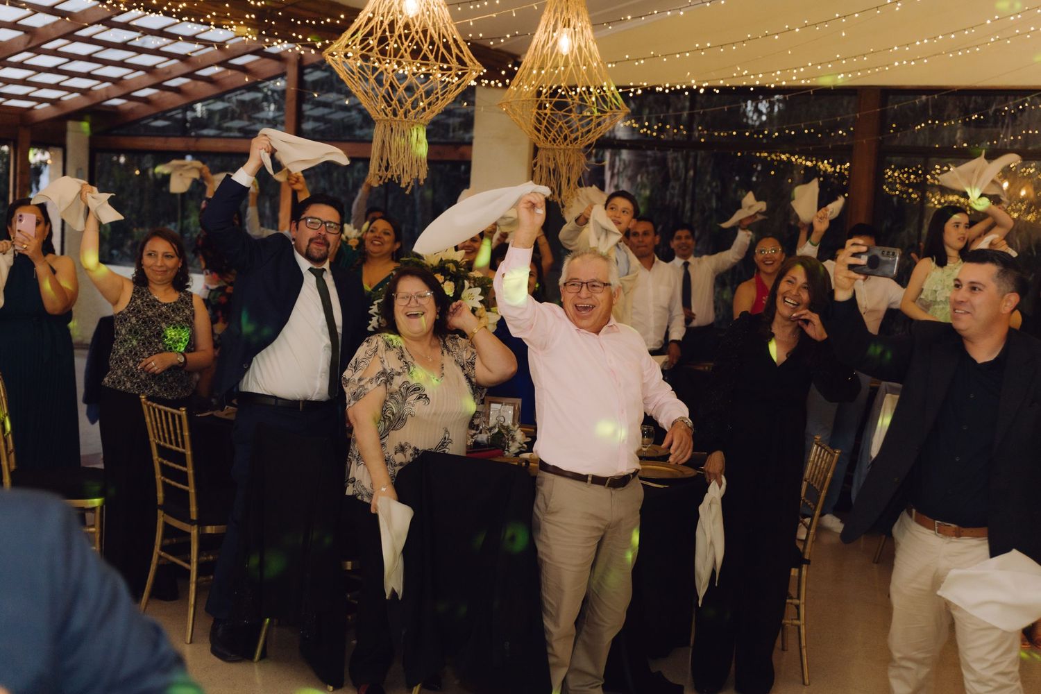 Excited wedding guests celebrating on the dance floor, waving napkins under elegant geometric chandeliers at a reception.