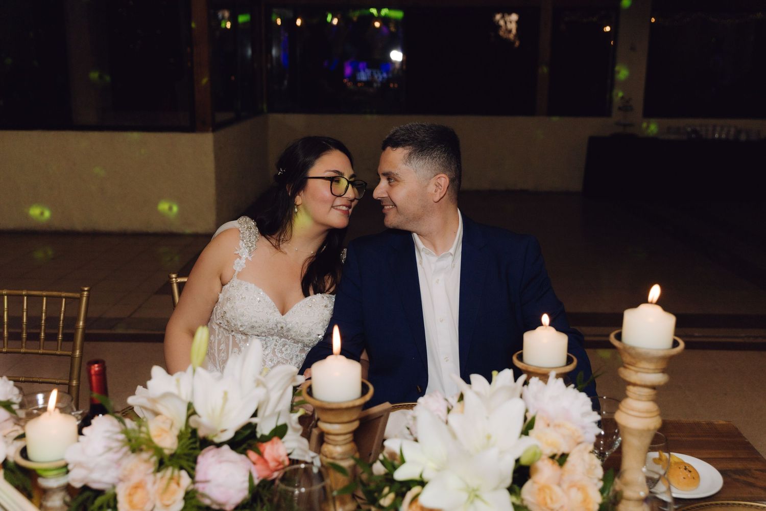 Bride in white lace gown and groom in navy suit share a romantic moment at candlelit sweetheart table with flowers.