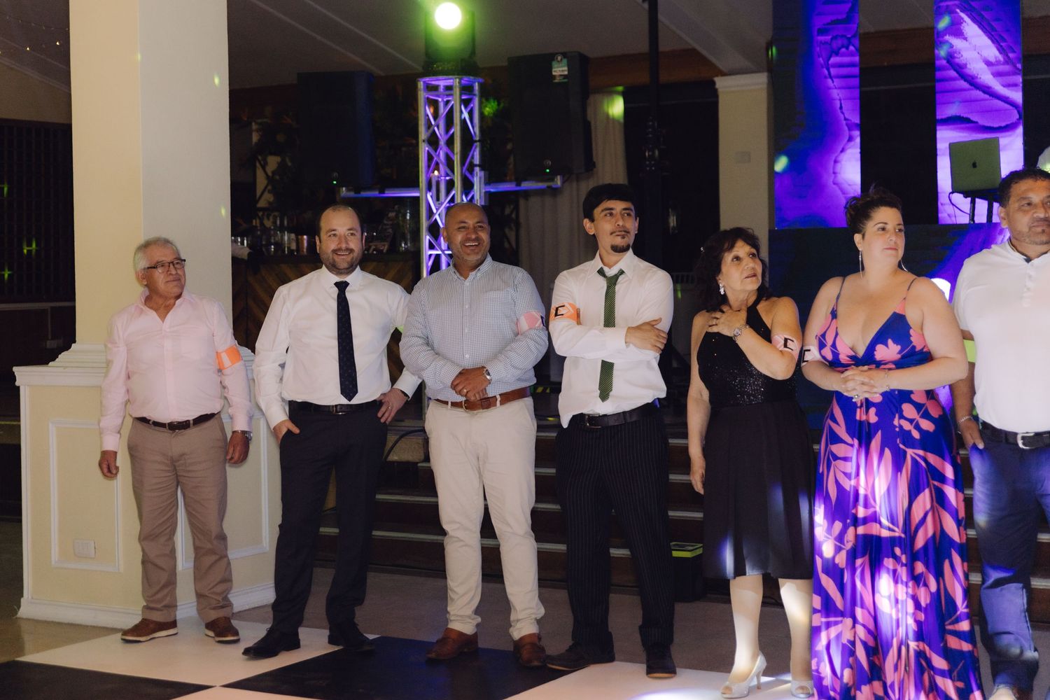 Smartly dressed wedding guests standing together on a dance floor beside a lit stage at an evening reception venue.