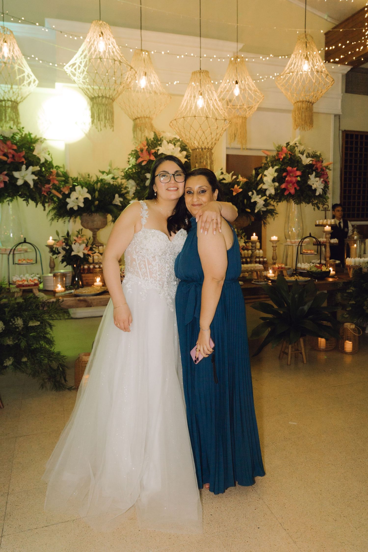 Bride in white tulle gown poses with a woman in teal dress in front of a floral arrangement and dessert table.