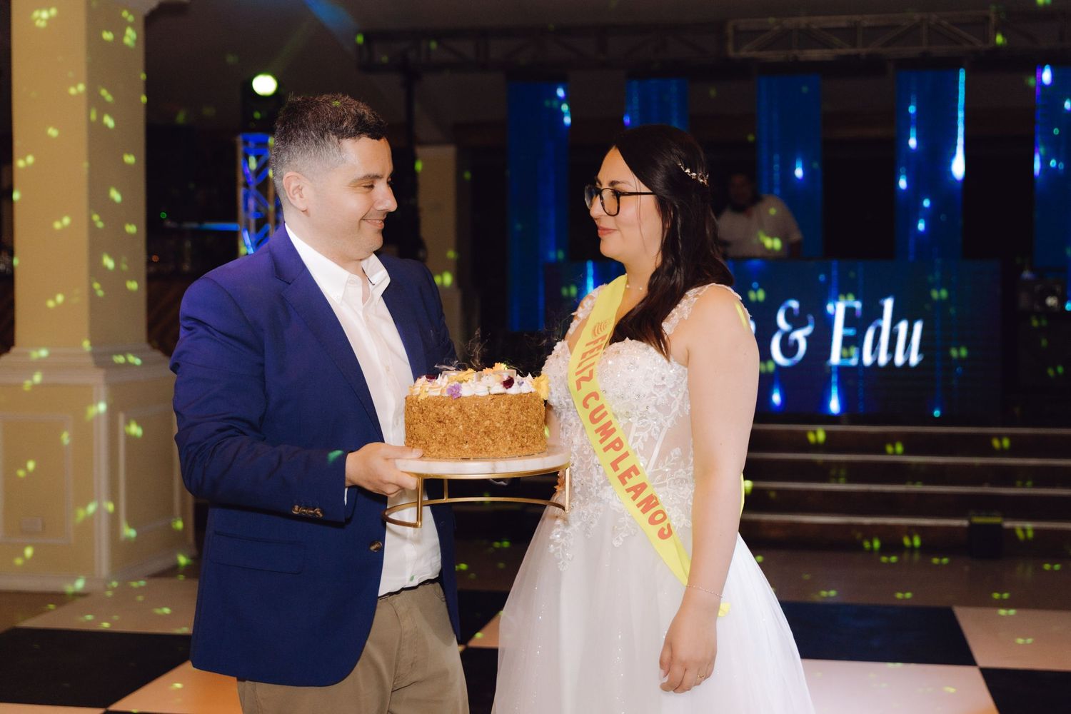 Groom in blue suit presents a cake to smiling bride wearing a yellow quinceañera sash at a wedding celebration.