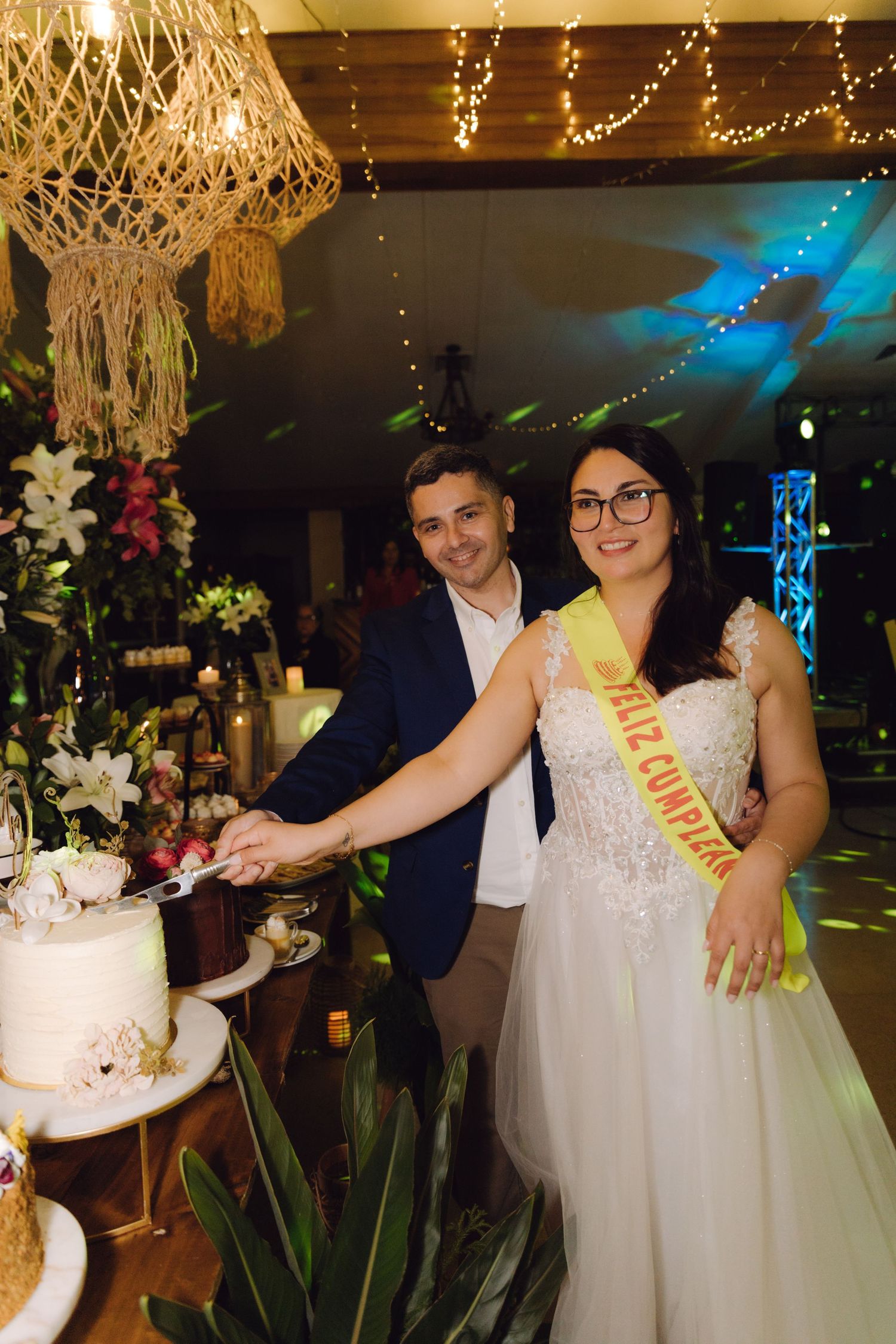 Bride and groom cut their white wedding cake together, bride wearing a yellow sash, surrounded by floral decor.