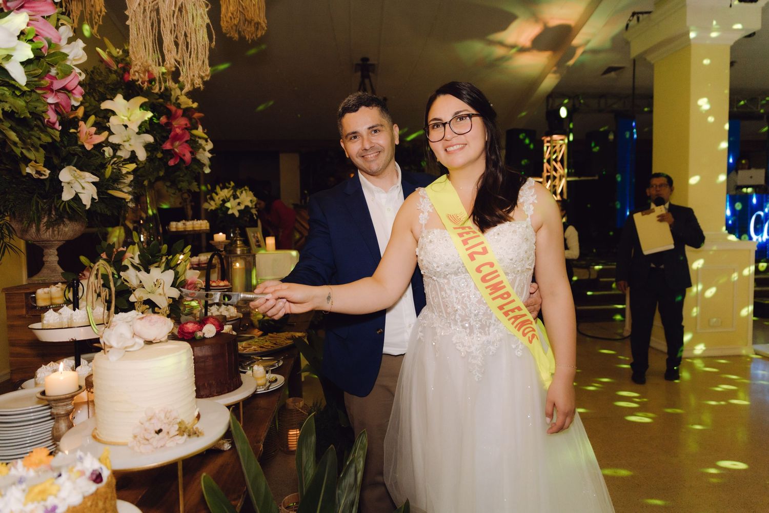 Smiling bride and groom pose beside their wedding cake, bride wearing a yellow celebratory sash at their reception.