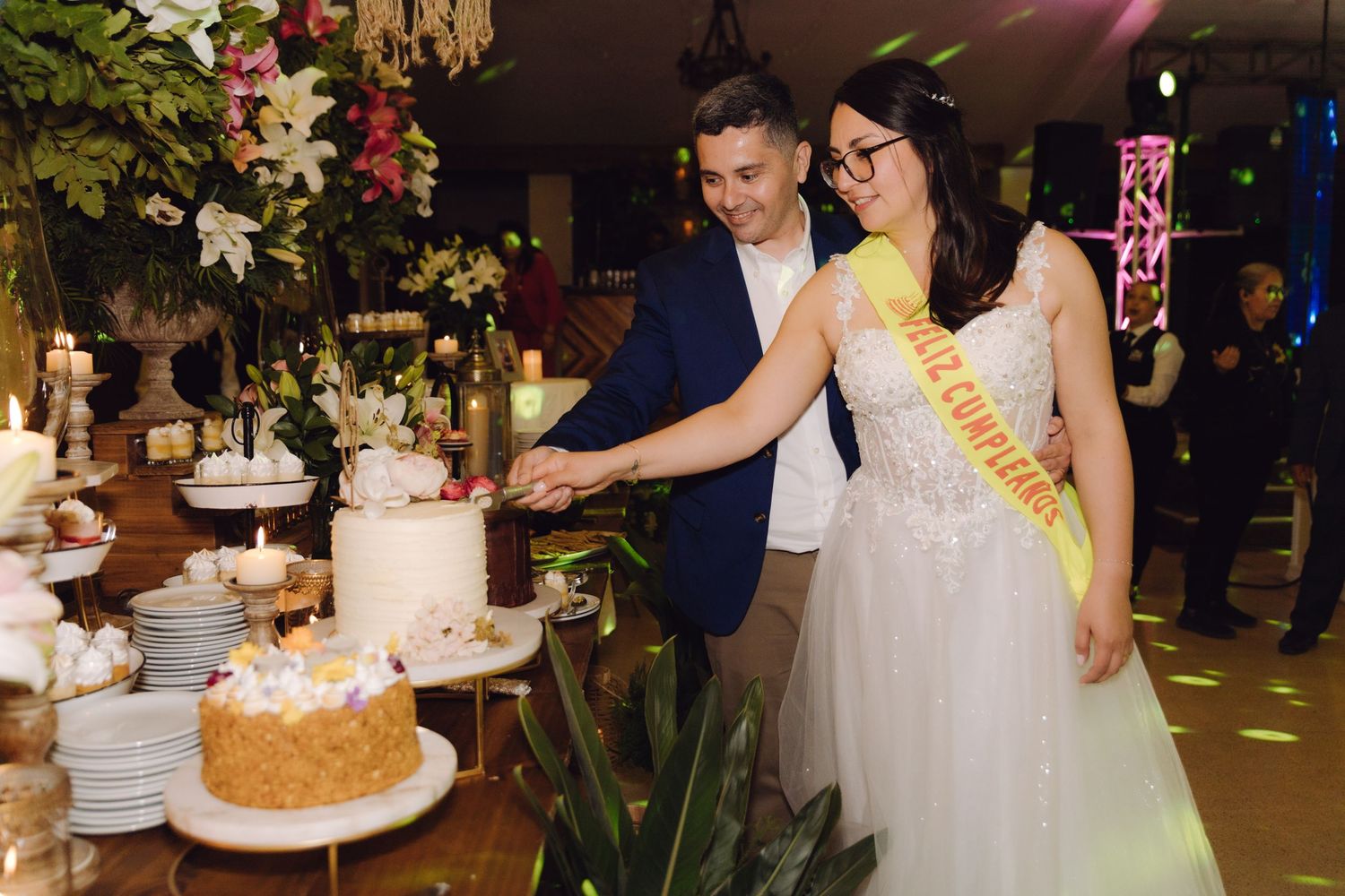 Bride and groom cutting wedding cake at elegant reception with floral decorations and candles.