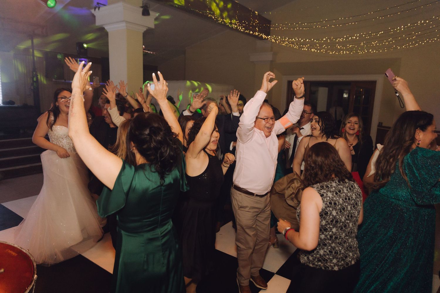 Wedding guests dancing with hands raised at lively indoor reception with fairy lights.