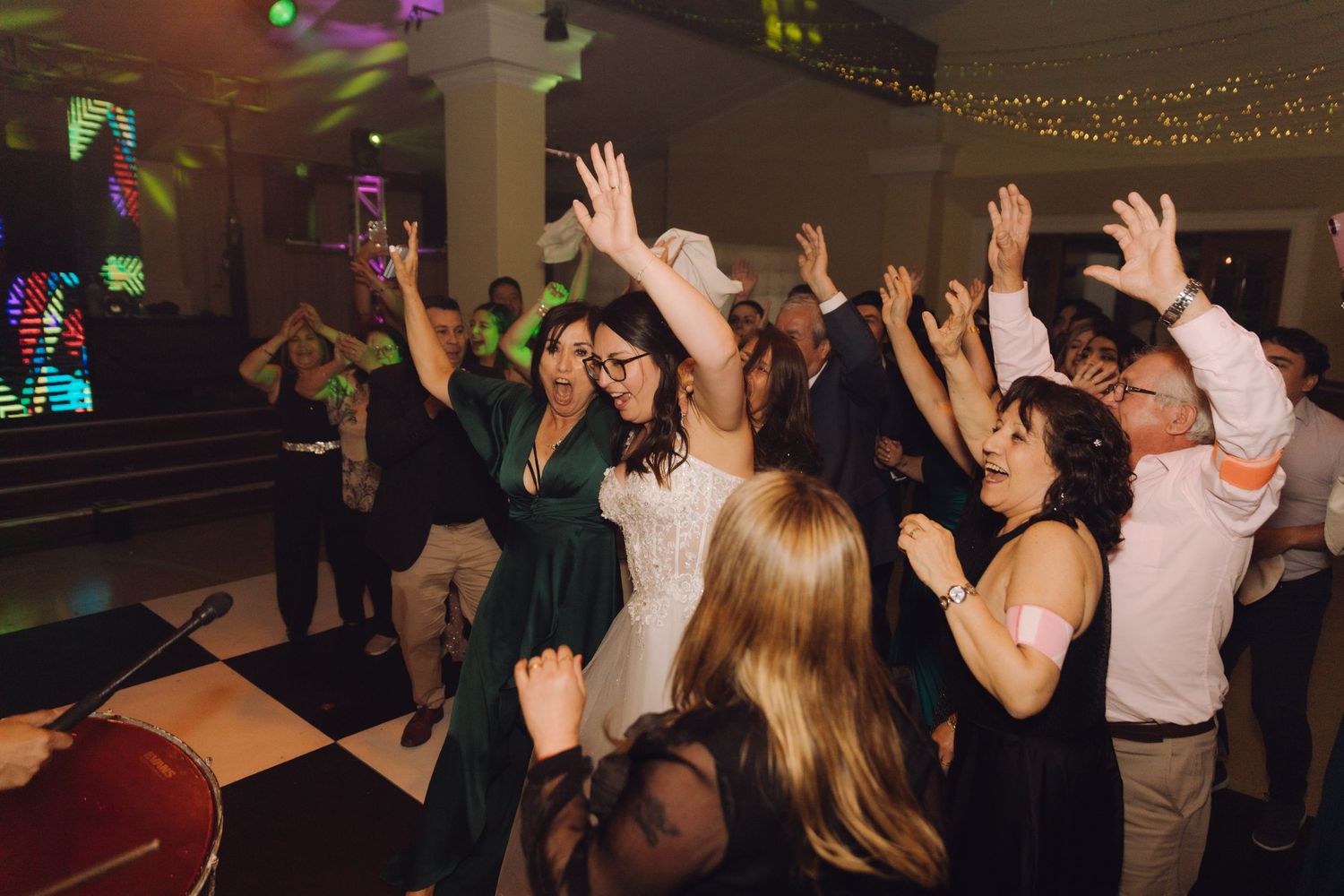 Bride and guests celebrating on dance floor with hands raised at wedding reception.