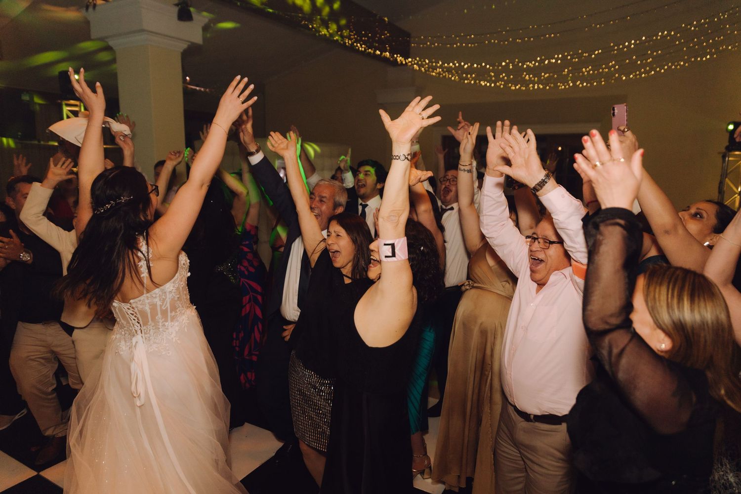 Joyful wedding guests dancing with arms raised under fairy lights at elegant reception.