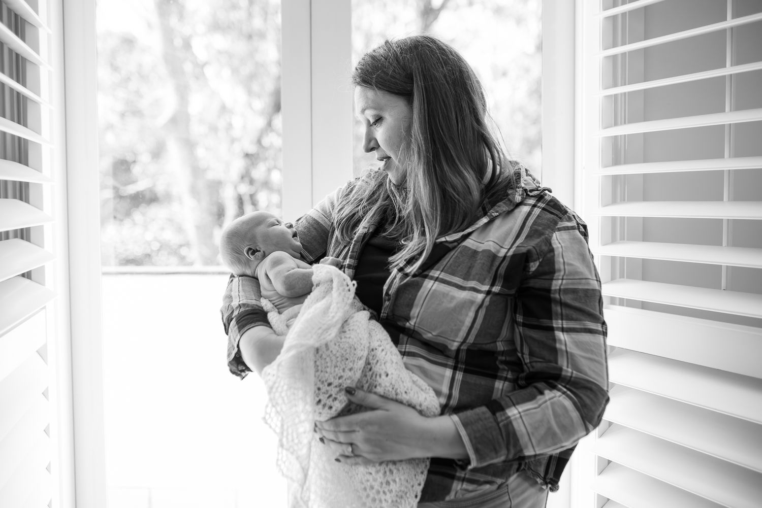 Black and white photo of a mother tenderly holding her newborn baby wrapped in a white blanket near a window in Melbourne.