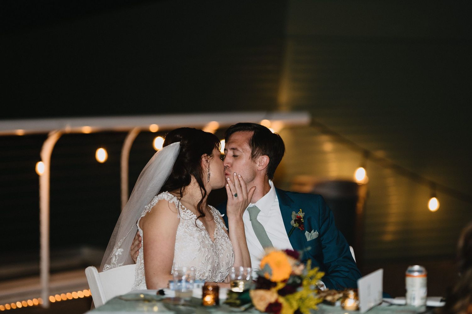 Bride and groom sharing a romantic kiss at their wedding reception table, surrounded by warm string lights and floral centerpieces.
