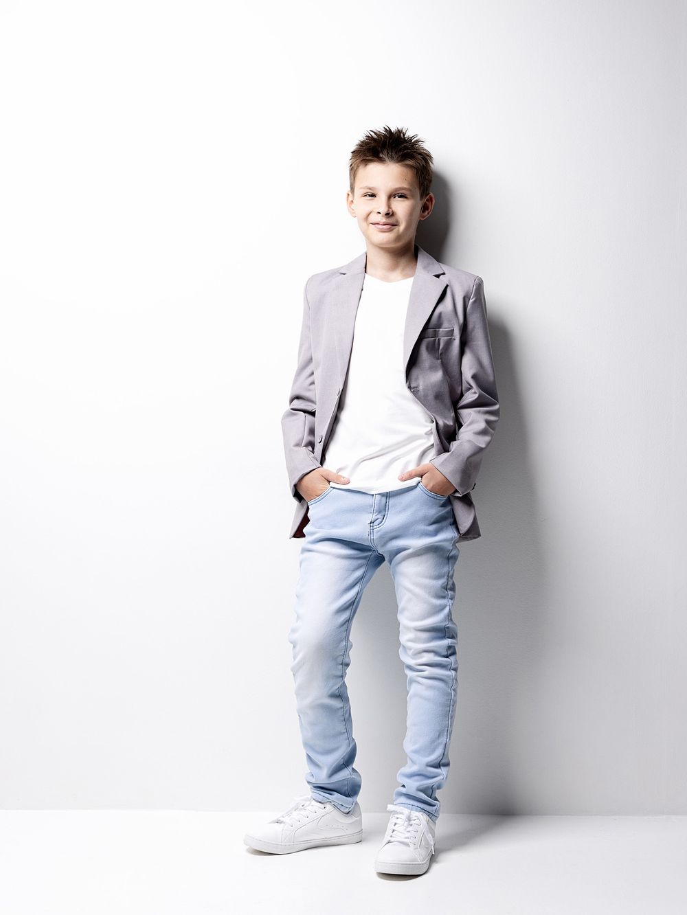 A young boy in a gray blazer, white tee, light jeans, and white sneakers poses confidently against a white wall.