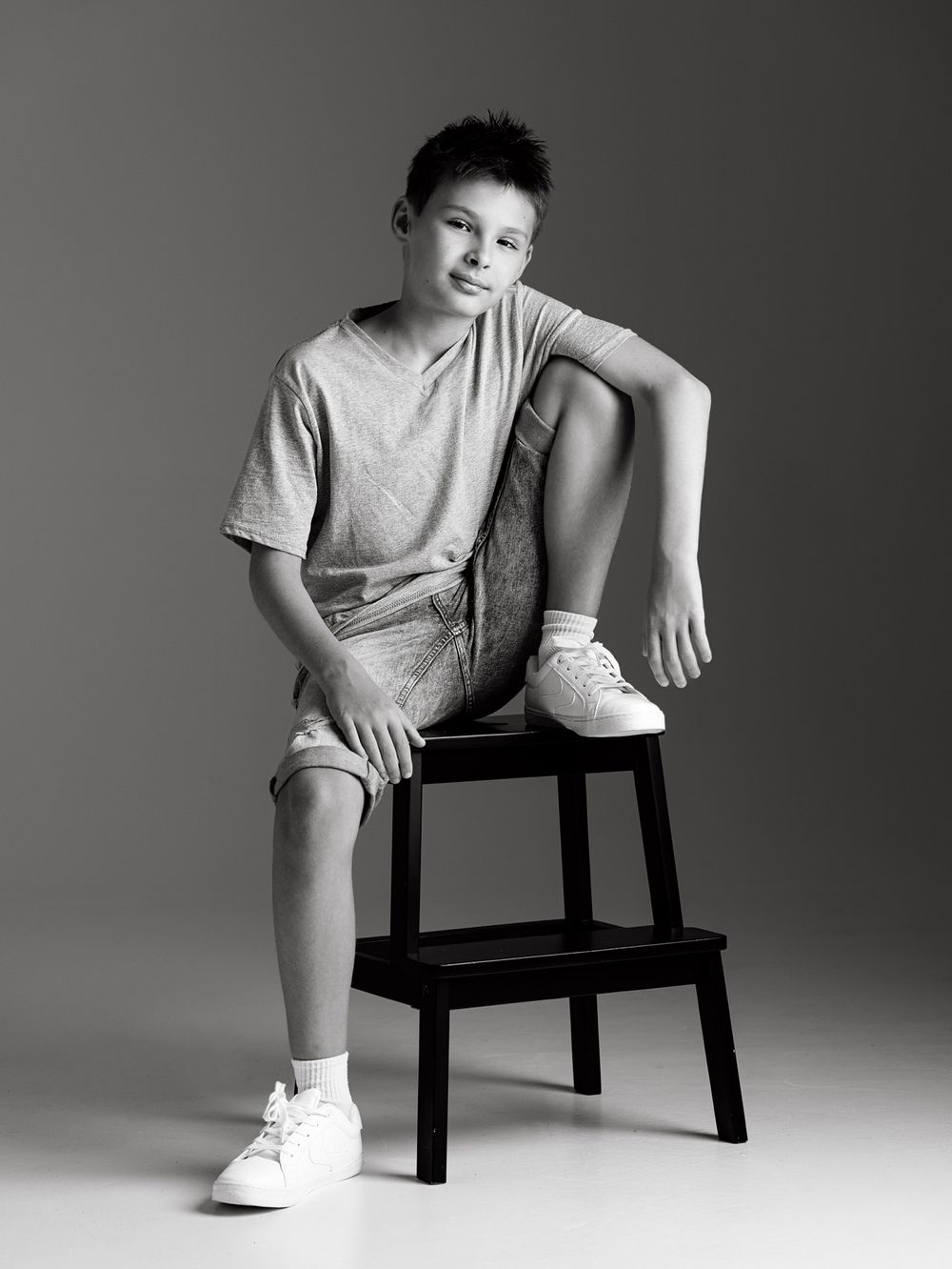 Black and white portrait of a young boy sitting on a stool in casual shorts and sneakers against a gray background.