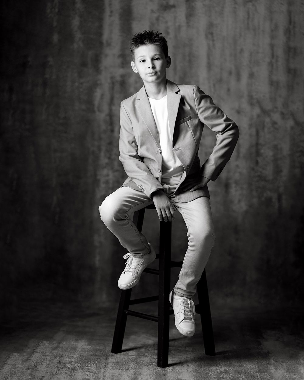 Black and white studio portrait of a young boy in a blazer and sneakers sitting confidently on a stool.