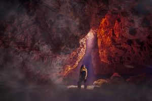 A lone woman stands in a glowing red cave as a shaft of purple light and mist illuminate the rugged rock walls around her.