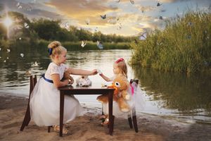 Two little girls in white dresses share a tea party at a small table on a lakeshore as butterflies fly through the golden light.