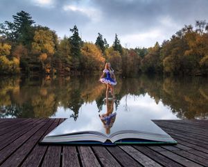 A ballerina in a colorful tutu appears to stand on an open book resting on a wooden dock overlooking a lake and autumn forest.