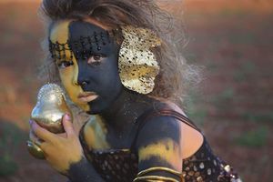 Girl with dark and gold face paint, a gold leaf ear accessory and gold bangles, holding a gilded pear in an artistic portrait.