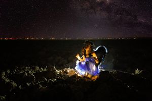 Girl in a glowing blue tutu crouches on dark rocks beneath a starry night sky with the Milky Way and distant city lights.