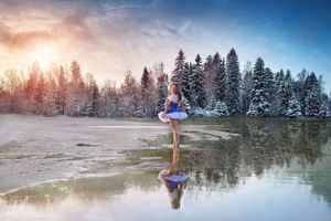 Ballerina in a pale blue tutu stands on a frozen lake at sunrise, reflected in the water in front of snowy evergreen trees.