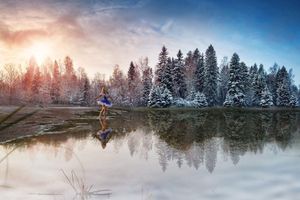 A ballerina in a blue tutu stands on a calm reflective lake before snow-dusted evergreen trees at a glowing winter sunrise.