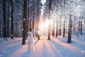 A woman in a white dress reaches toward glowing sunlight streaming through snowy pine trees as snowflakes fall in a winter forest.