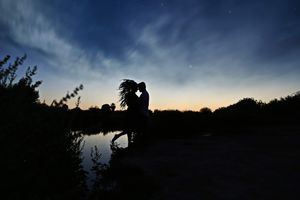 A couple stands embracing in silhouette beside a still lake under a deep blue twilight sky with soft clouds and a faint glow on the horizon.