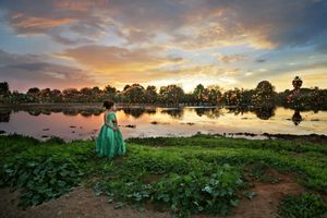 A woman in a flowing emerald green gown stands beside a reflective lake under a dramatic golden and purple sunset sky with distant trees.