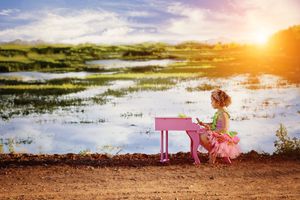 A little girl in a pink tutu plays a small pink toy piano beside a shimmering wetland at sunset on a dirt path.