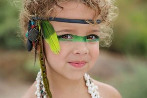Smiling young child with green face paint, a feather headband and a white shell necklace posing outdoors in a bohemian-style portrait.
