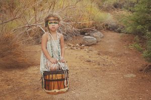 Child in a light fringed dress and green face paint sits behind a handmade drum on a dirt trail surrounded by dry desert brush.