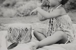 Black and white photo of a child in a fringed top and beaded necklace sitting on a blanket beside a decorated drum and a feather.