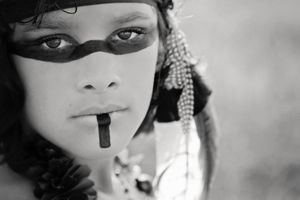 Black and white close-up portrait of a child with dark face paint across the eyes, beaded accessories and an intense, steady gaze.