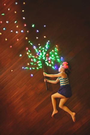 Girl lying on a wooden floor holding a branch of glowing multicolored fairy lights, with light sparkles drifting across the floor.
