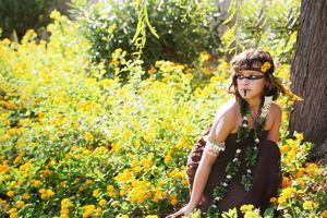 Child with a feather headband and green beaded necklaces crouches in a field of yellow flowers beside a tree trunk in warm sunlight.