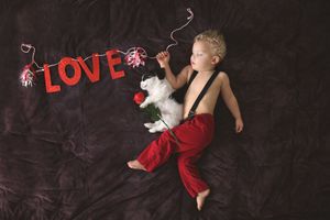 Toddler in red pants and suspenders lies beside a white puppy and red rose under a red LOVE banner on a dark fabric backdrop.