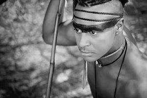 Black and white portrait of a young man with striped face paint, a headband and a necklace, holding a spear and gazing intently.