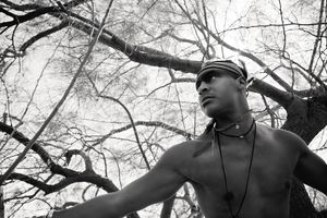 Black and white photo of a shirtless young man in a headband and beaded necklace stretching his arm beneath bare tree branches.